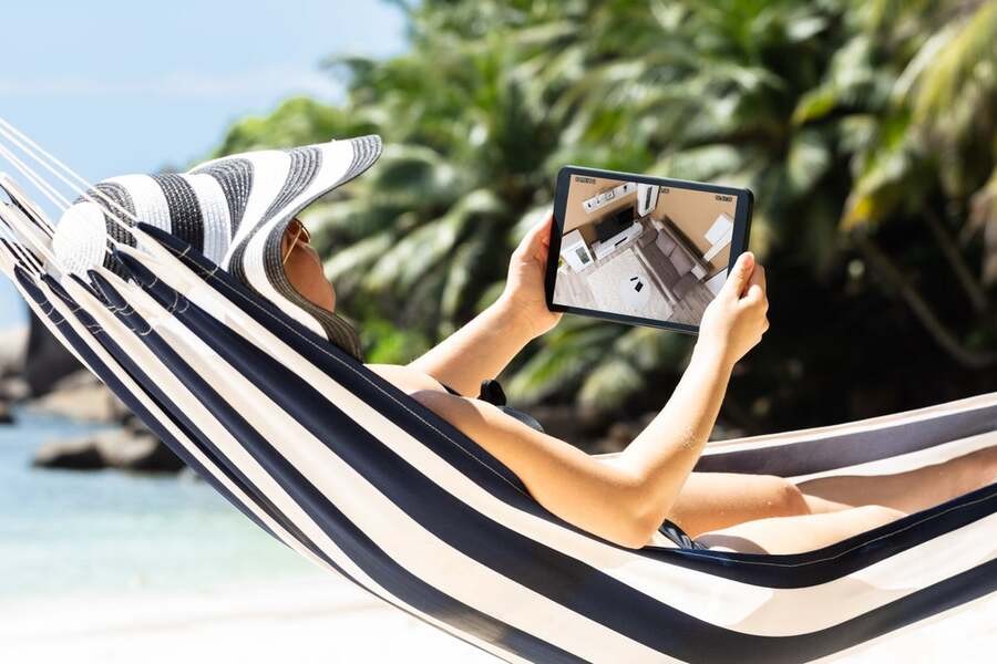 A woman relaxes on a hammock on the beach while checking on her home through her tablet.