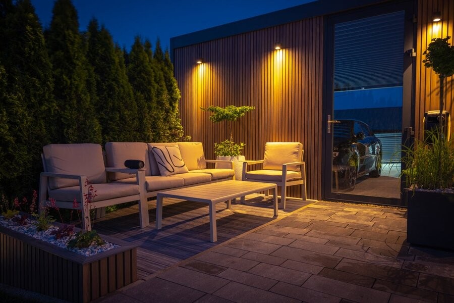 An outdoor patio area in a backyard at night, illuminated by outdoor lighting fixtures.